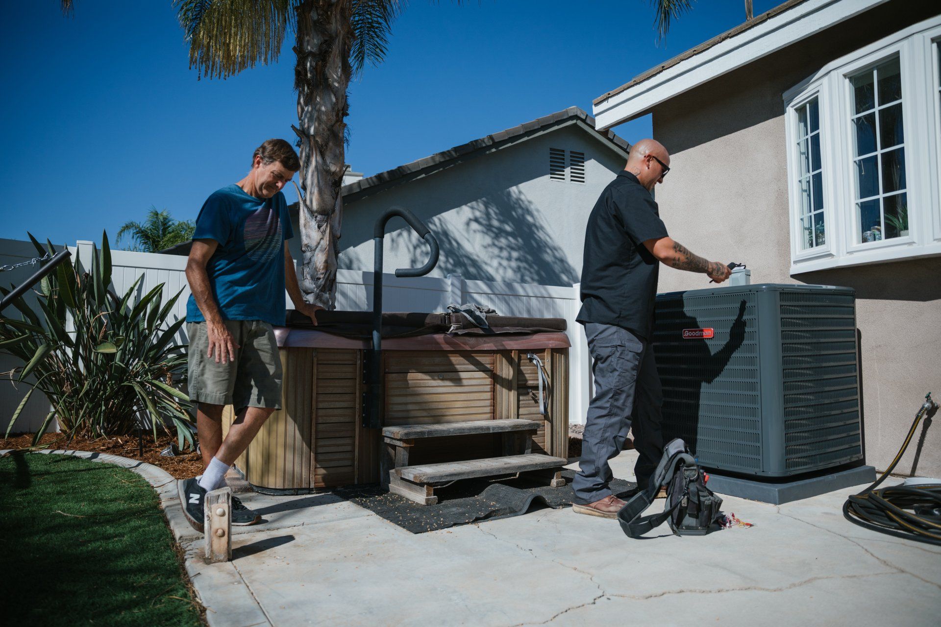 Two men near an outdoor AC unit and hot tub on a sunny day. One works on the unit, the other watches.