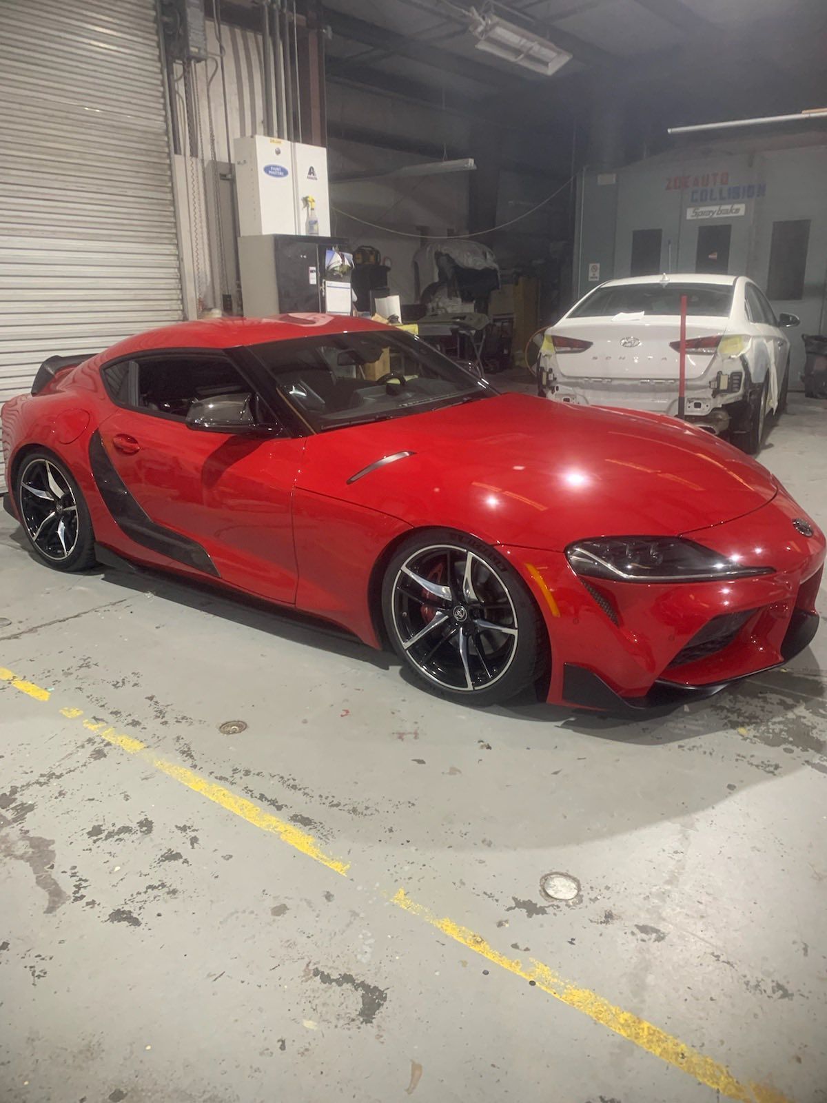 Red Toyota Supra sports car in a garage with a partially repaired white car in the background.