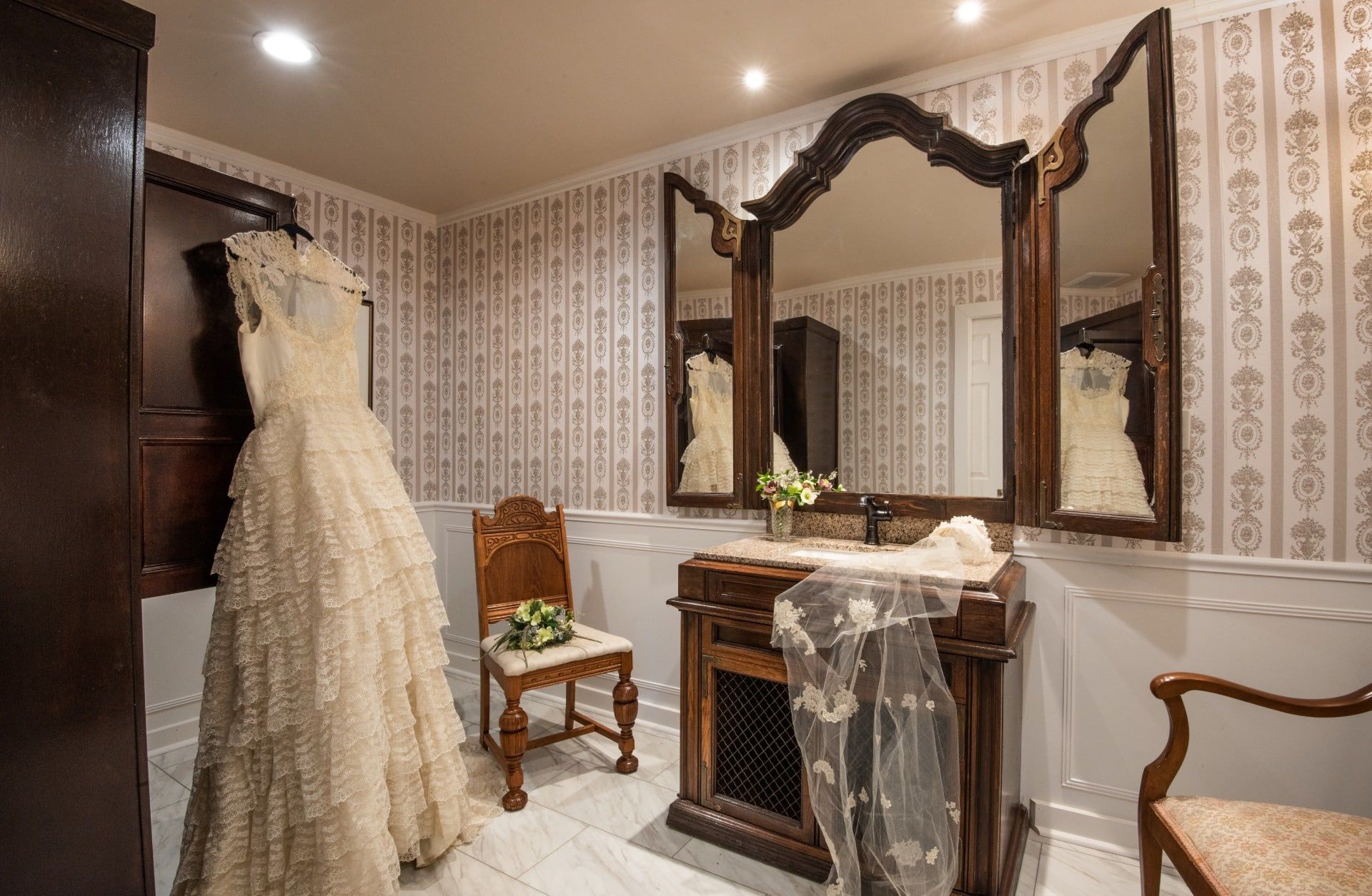 A wedding dress is hanging in a bathroom next to a chair.