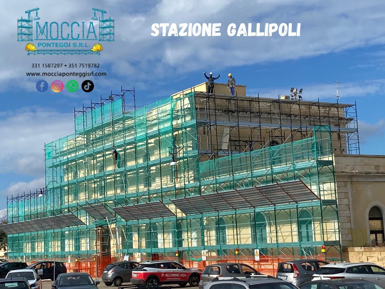 Ponteggi con rete verde ricoprono la stazione ferroviaria di Gallipoli mentre gli operai edili effettuano lavori di riparazione esterna.