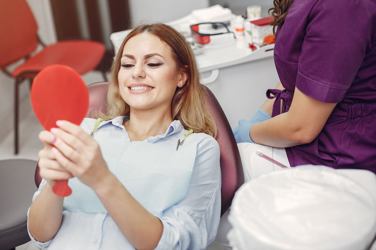 A woman is sitting in a dental chair looking at her teeth in a mirror.