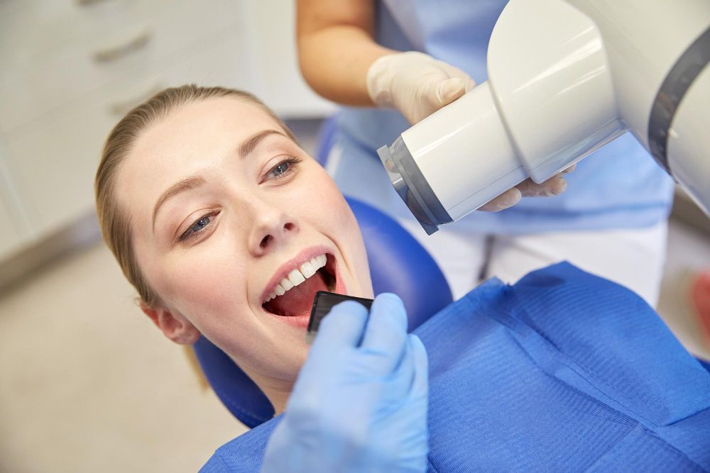 Woman getting dental X-ray. Dentist in gloves holding device near her open mouth. Blue chair and drape.