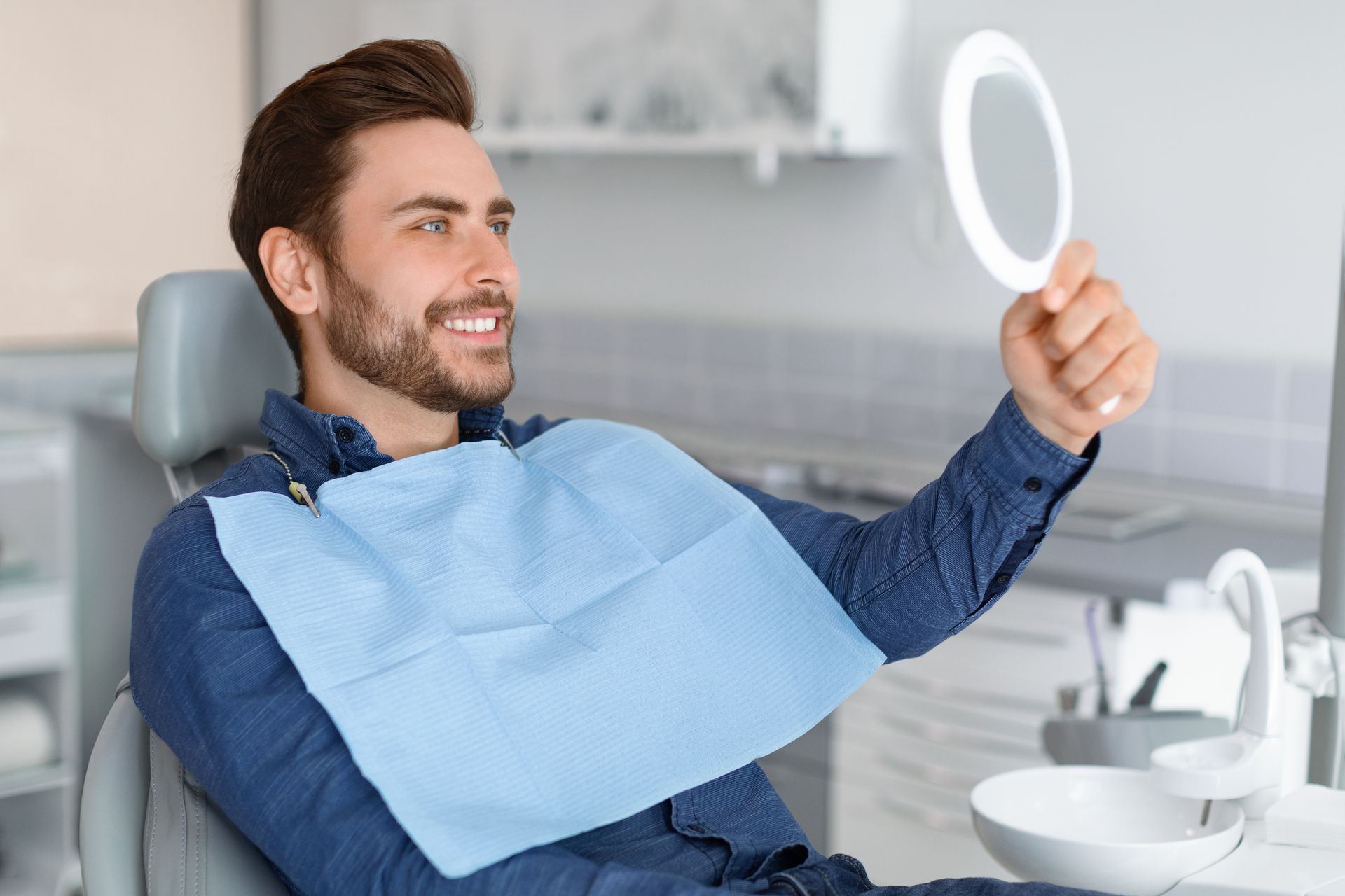 A man is sitting in a dental chair looking at his teeth in a mirror.