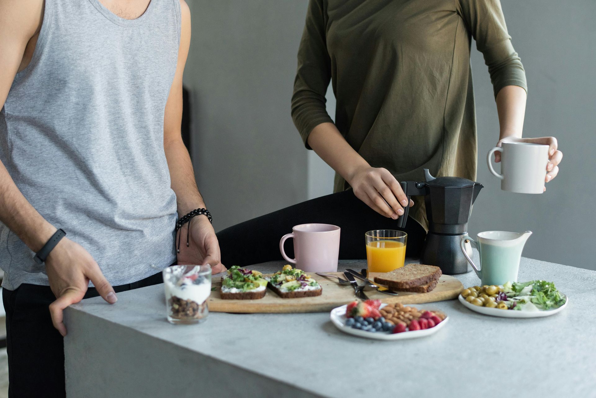 Two people preparing a meal of bread, fruit, and vegetables with multiple mugs of drinks on a table