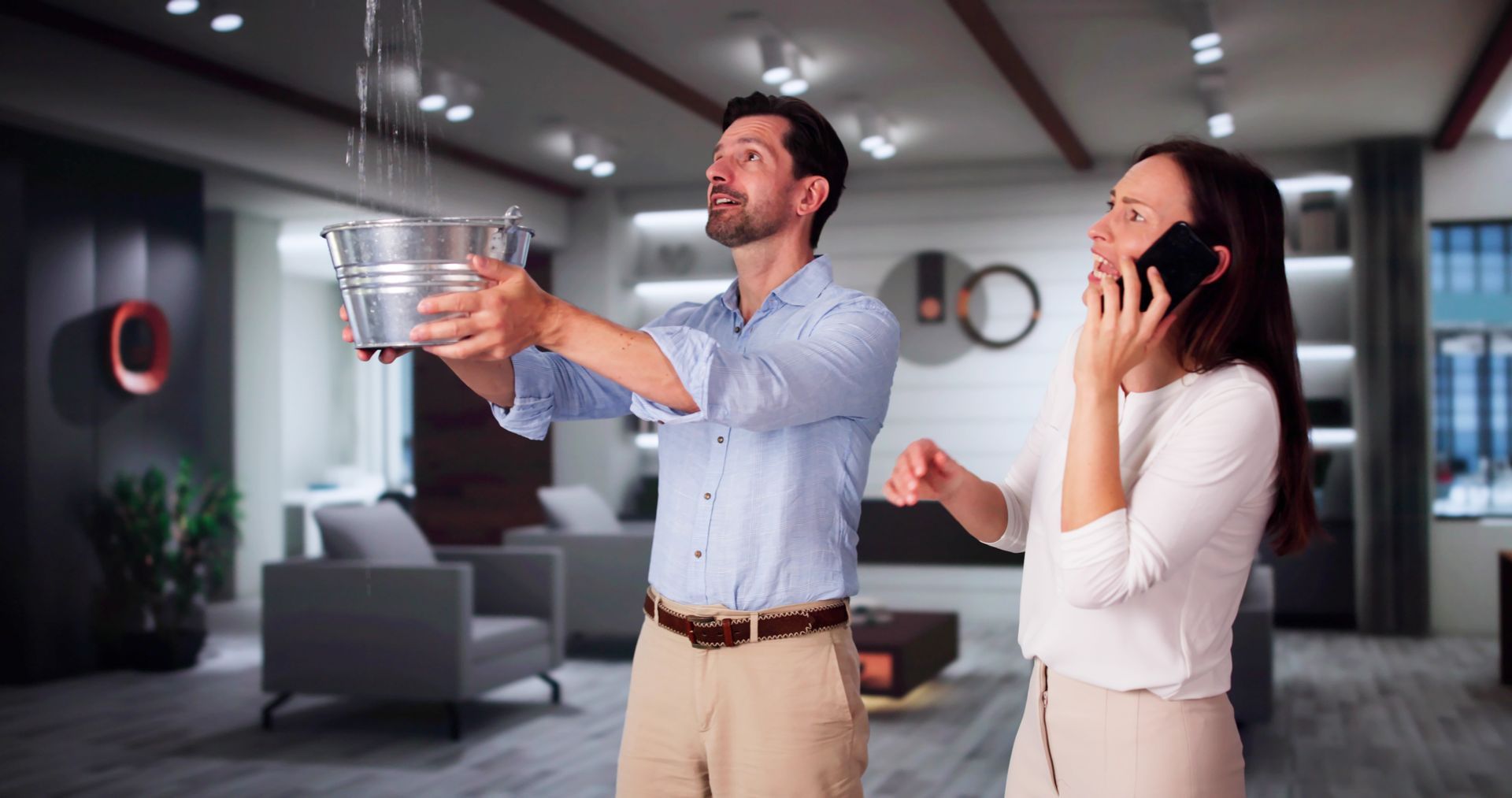 Man holding a bucket catching water from a ceiling leak, woman on the phone in a modern living room.