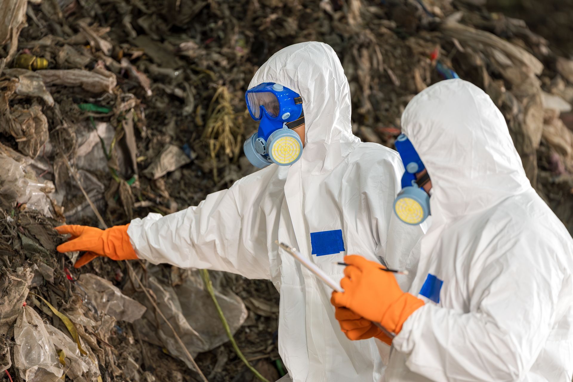 Two people in white hazmat suits examine a landfill, one pointing, both wearing respirators.