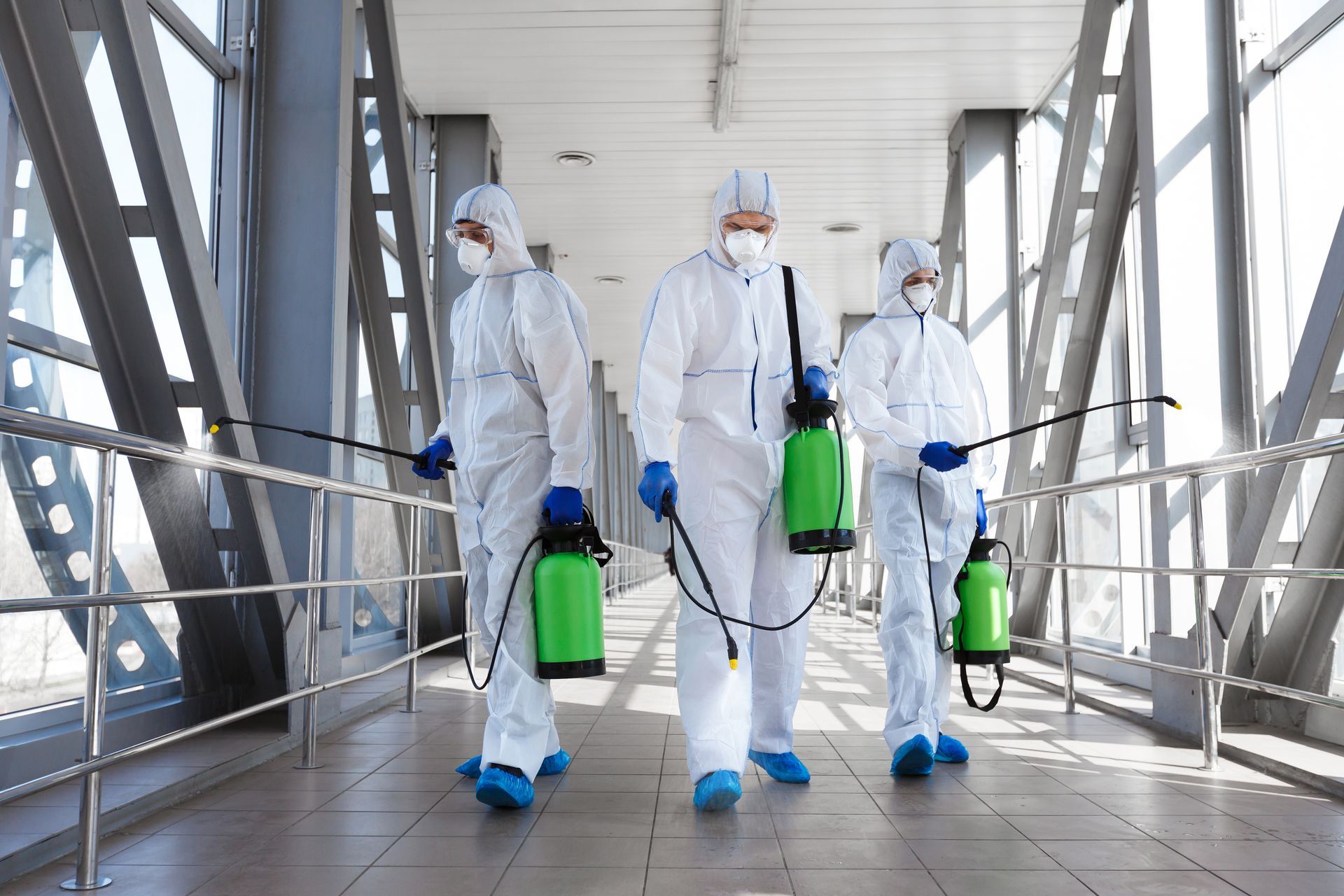 Three people in white hazmat suits spraying disinfectant in a brightly lit corridor.