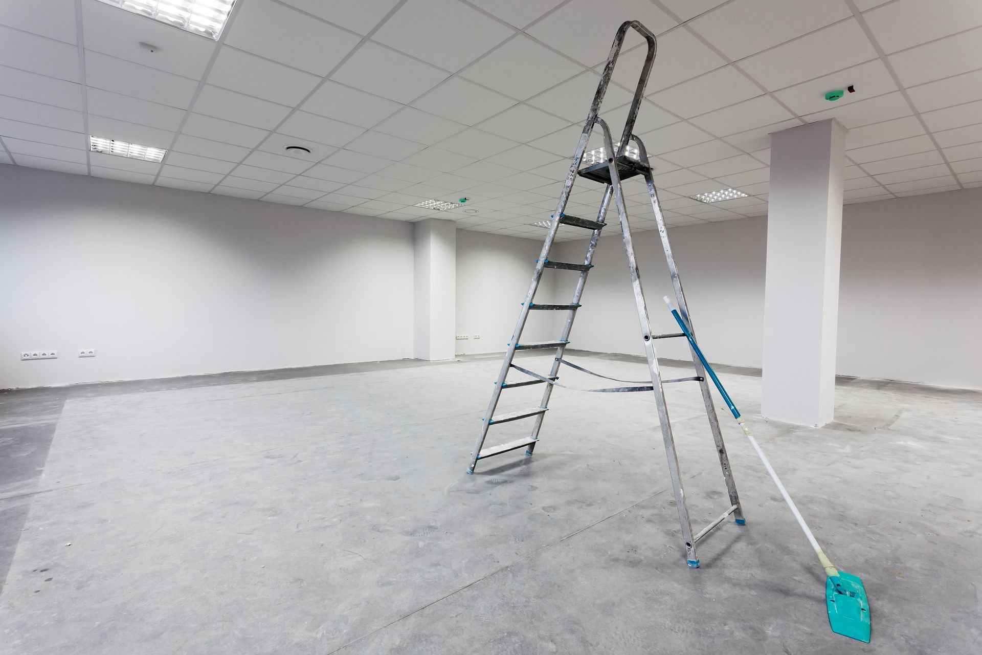 A ladder and mop in an empty room with white walls and concrete floor.