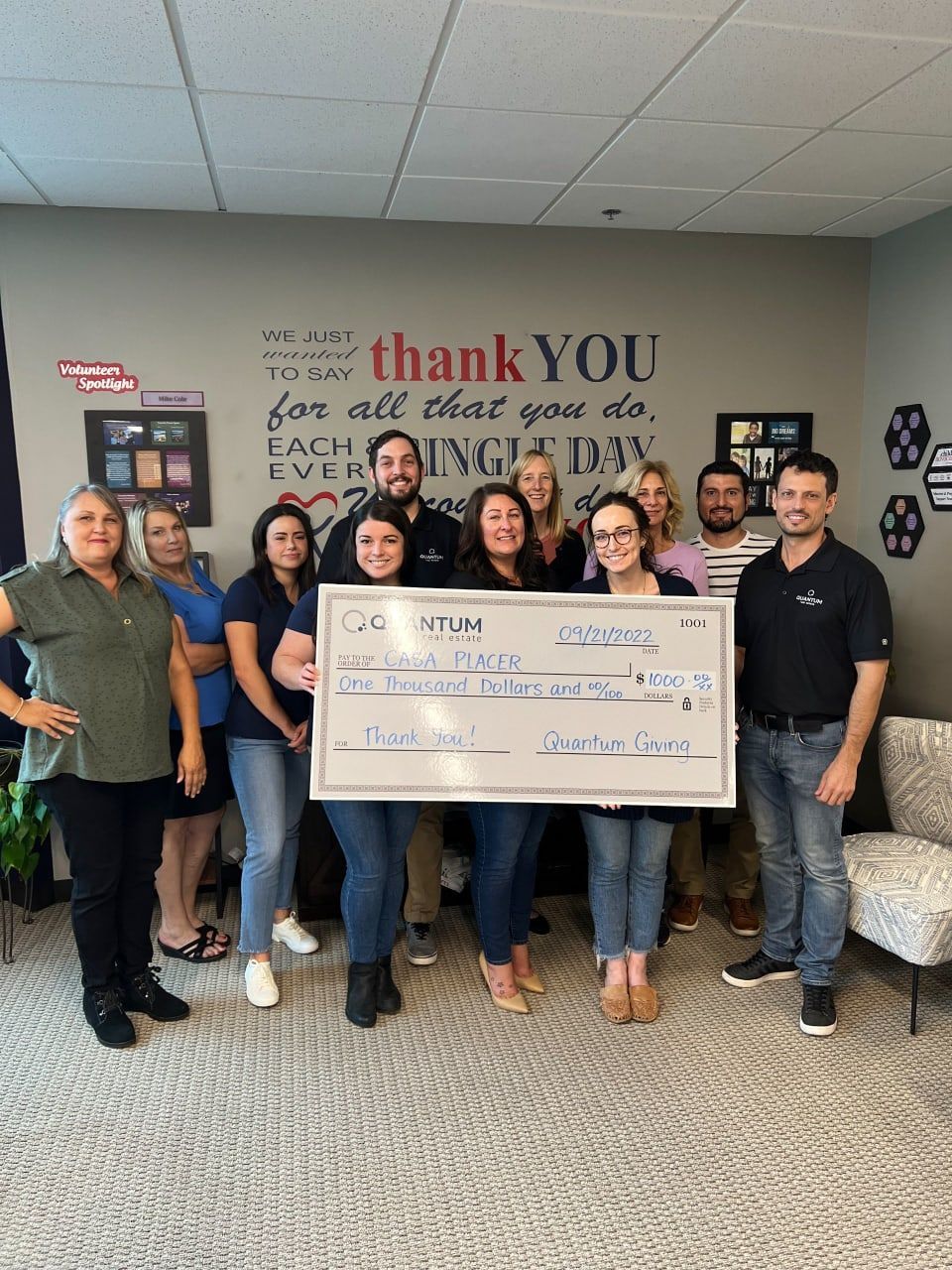 Group of people holding an oversized check, smiling in an office. Gray wall with