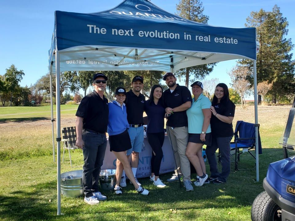 Group of eight people at an outdoor real estate event, posing in front of a branded tent.