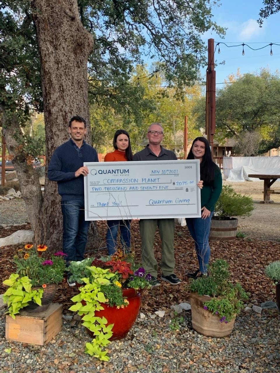 Four people hold a large check outside. Trees and foliage surround them; they are smiling.