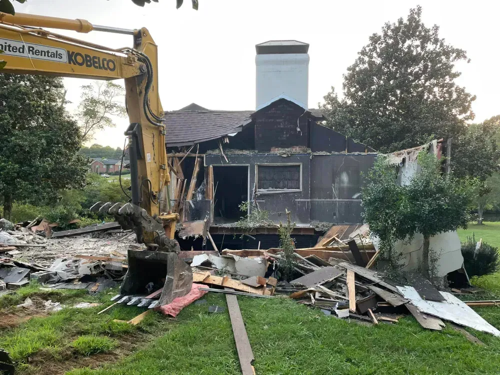 A house is being demolished by a yellow excavator.