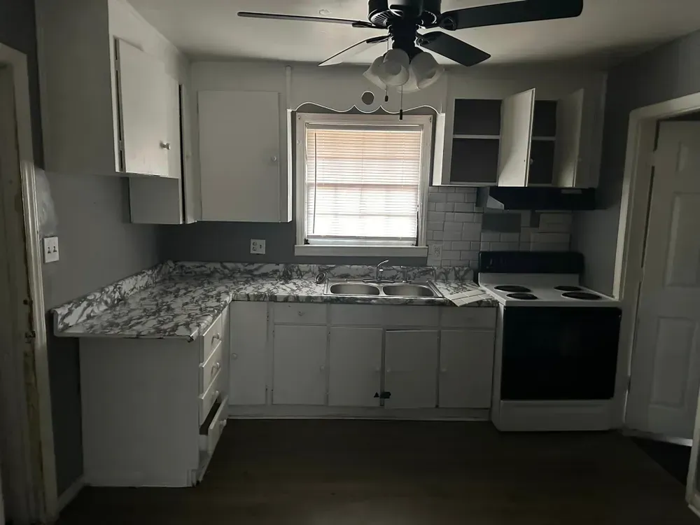 A kitchen with white cabinets , a stove , a sink and a ceiling fan.