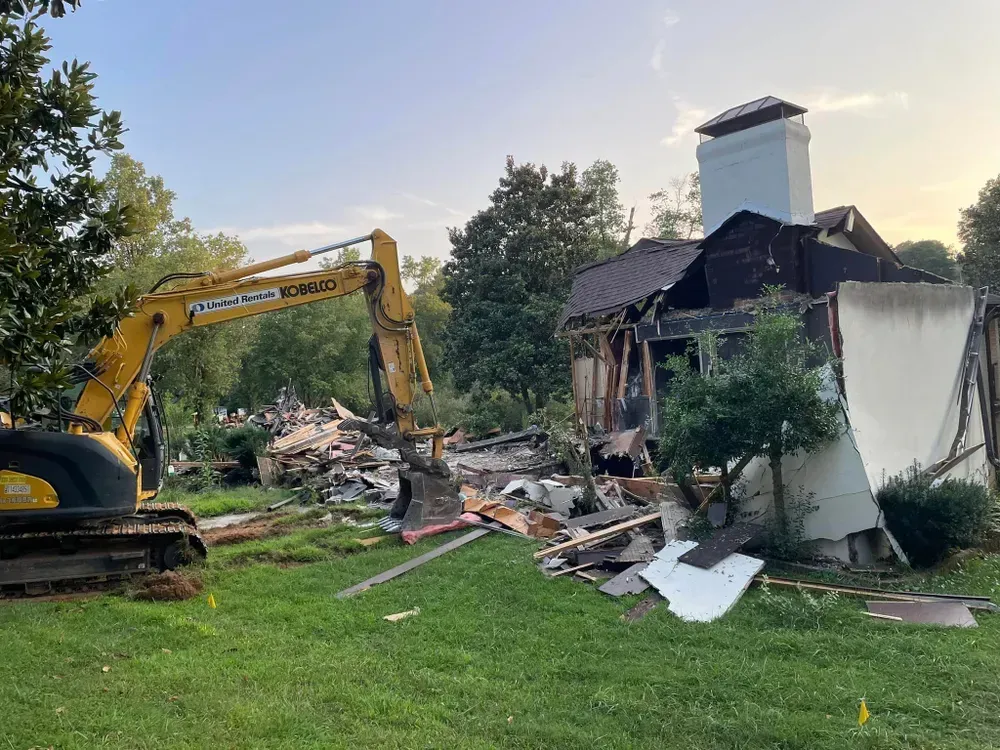 A yellow excavator is demolishing a house in a grassy field.