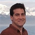 A person with dark hair and a neutral expression, posing in front of a body of water and snow-capped mountains.