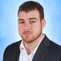 A person with short brown hair and a beard, wearing a white shirt and dark blazer against a blue backdrop.