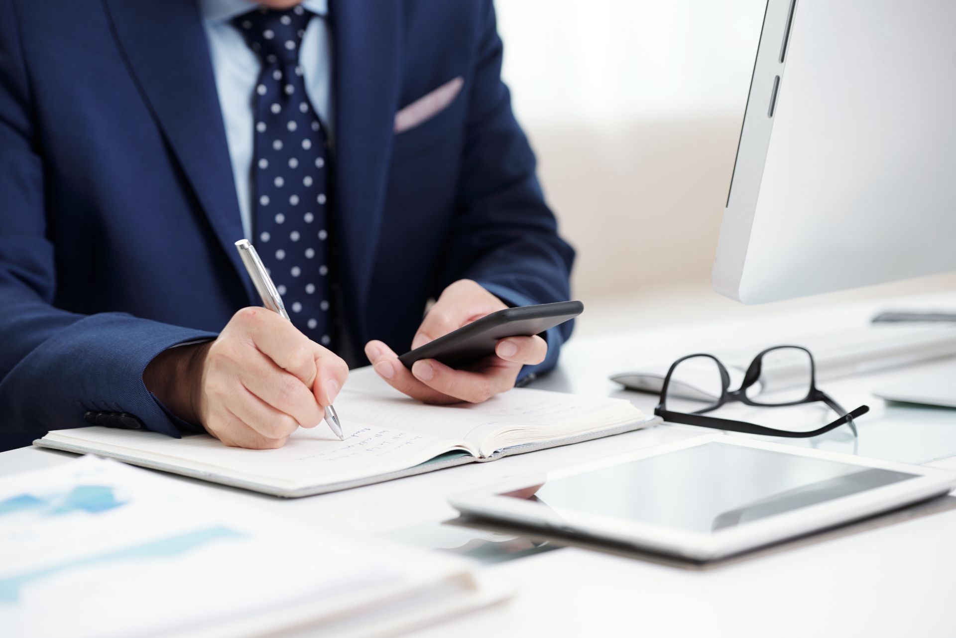 A person in a blue suit and polka-dot tie writing in a notebook while holding a smartphone at a desk with a tablet.