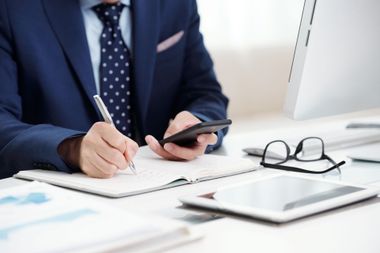 A person in a blue suit and polka-dot tie writes in a notebook while holding a smartphone at a desk with a tablet.