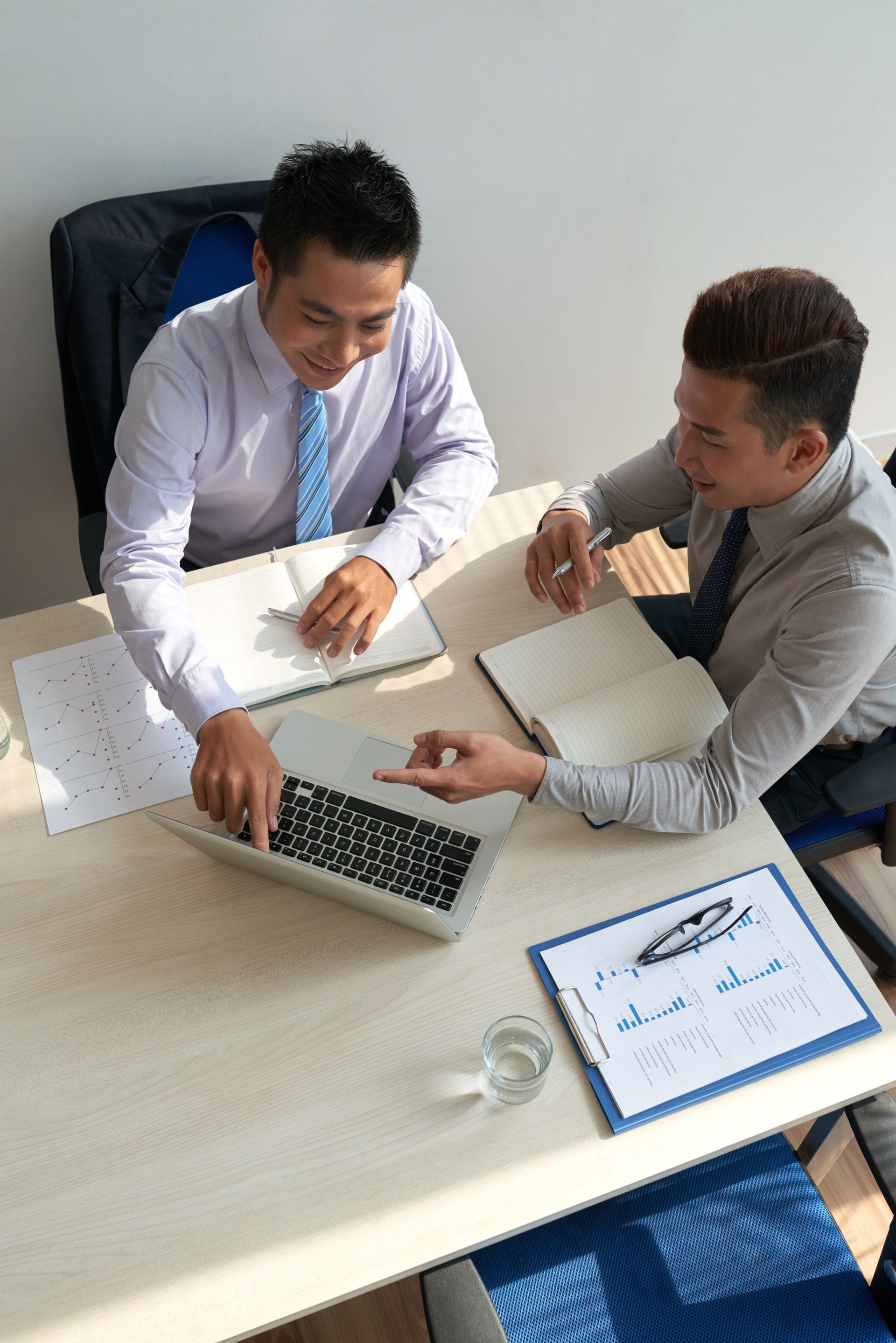 Two professionals in formal attire sit at a table collaborating on a laptop and documents in a bright office space.