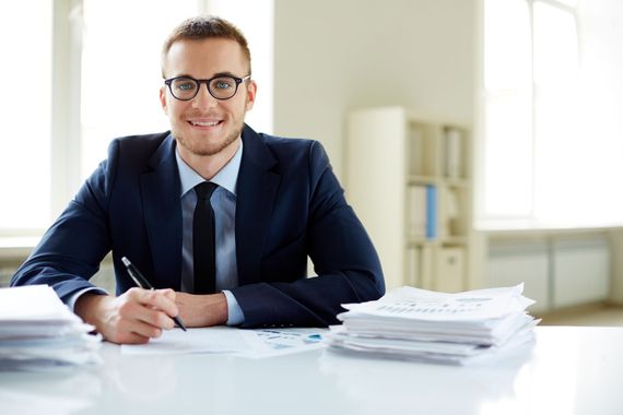 A smiling professional in a suit and glasses sitting at a desk with stacks of documents, holding a pen.