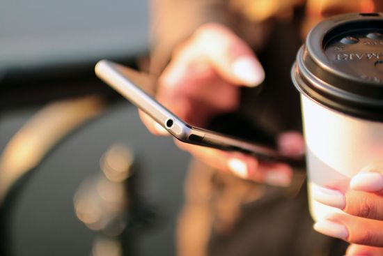 Close-up of hands holding a smartphone and a takeaway coffee cup against a softly blurred, warm outdoor background.
