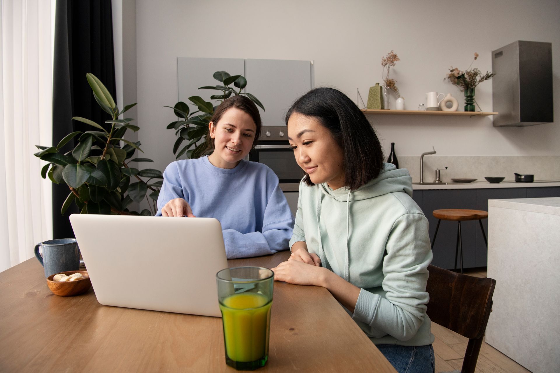 Two people sit at a wooden table in a bright kitchen, looking at a laptop together while one points at the screen.