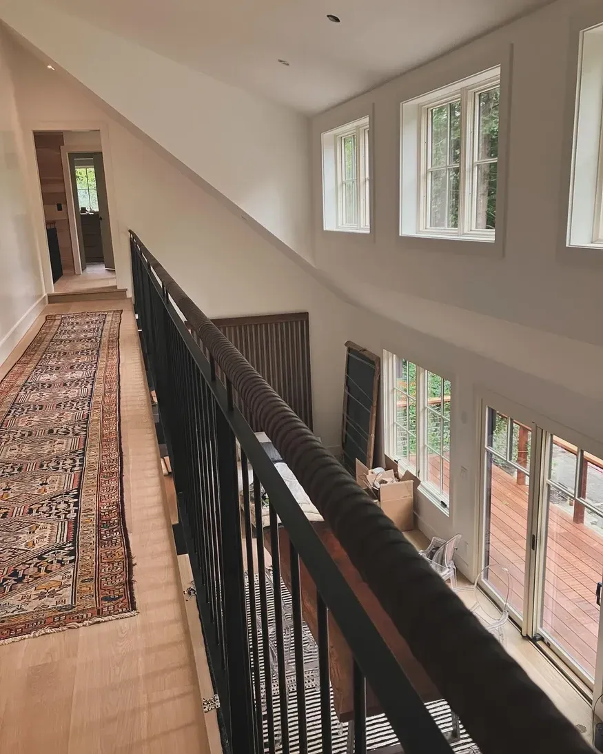 Interior shot of a hallway and staircase with dark railing, light walls, windows.