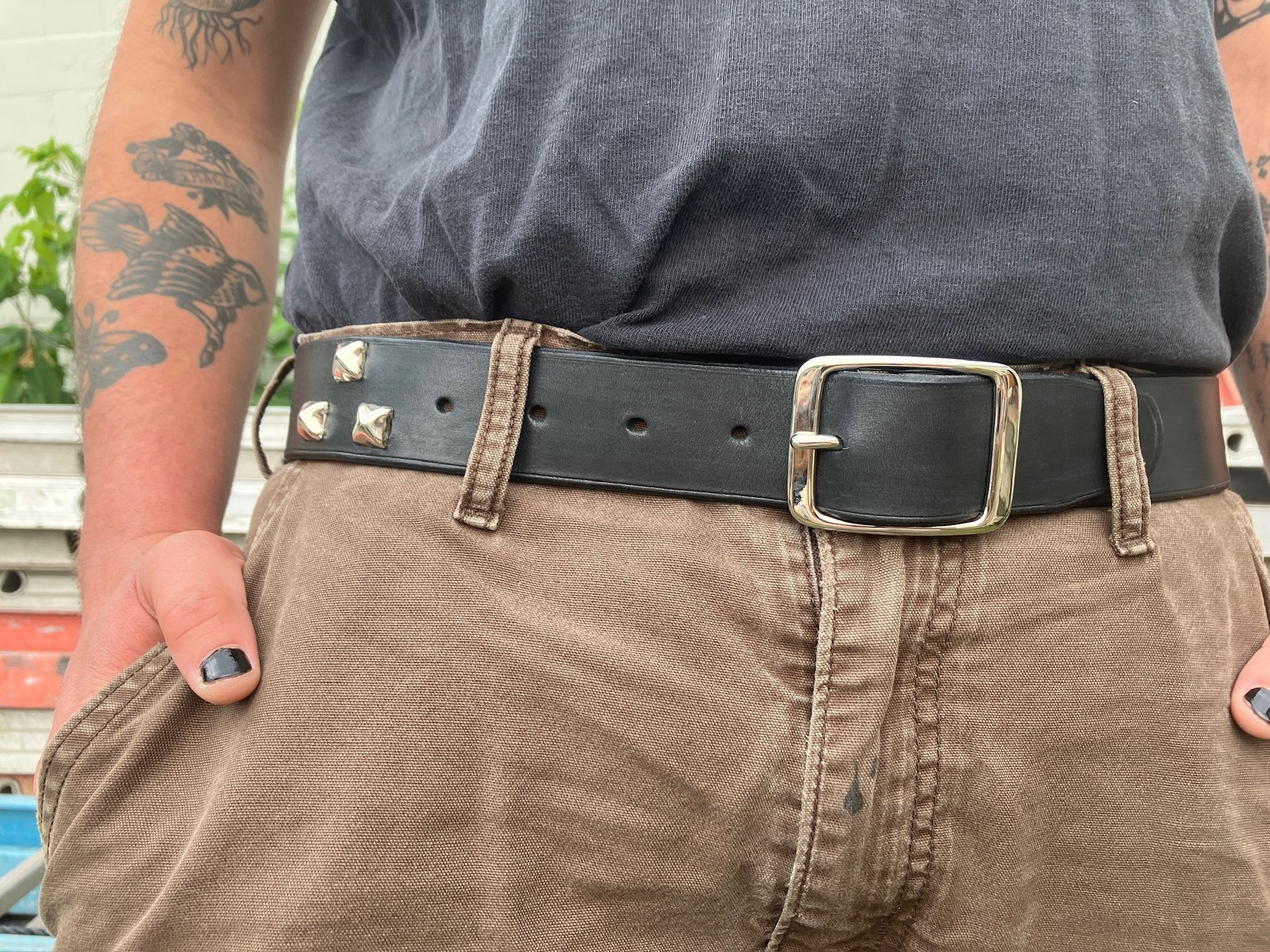 Person wearing a studded black belt over brown pants, hands in pockets. Tattoo on left arm.