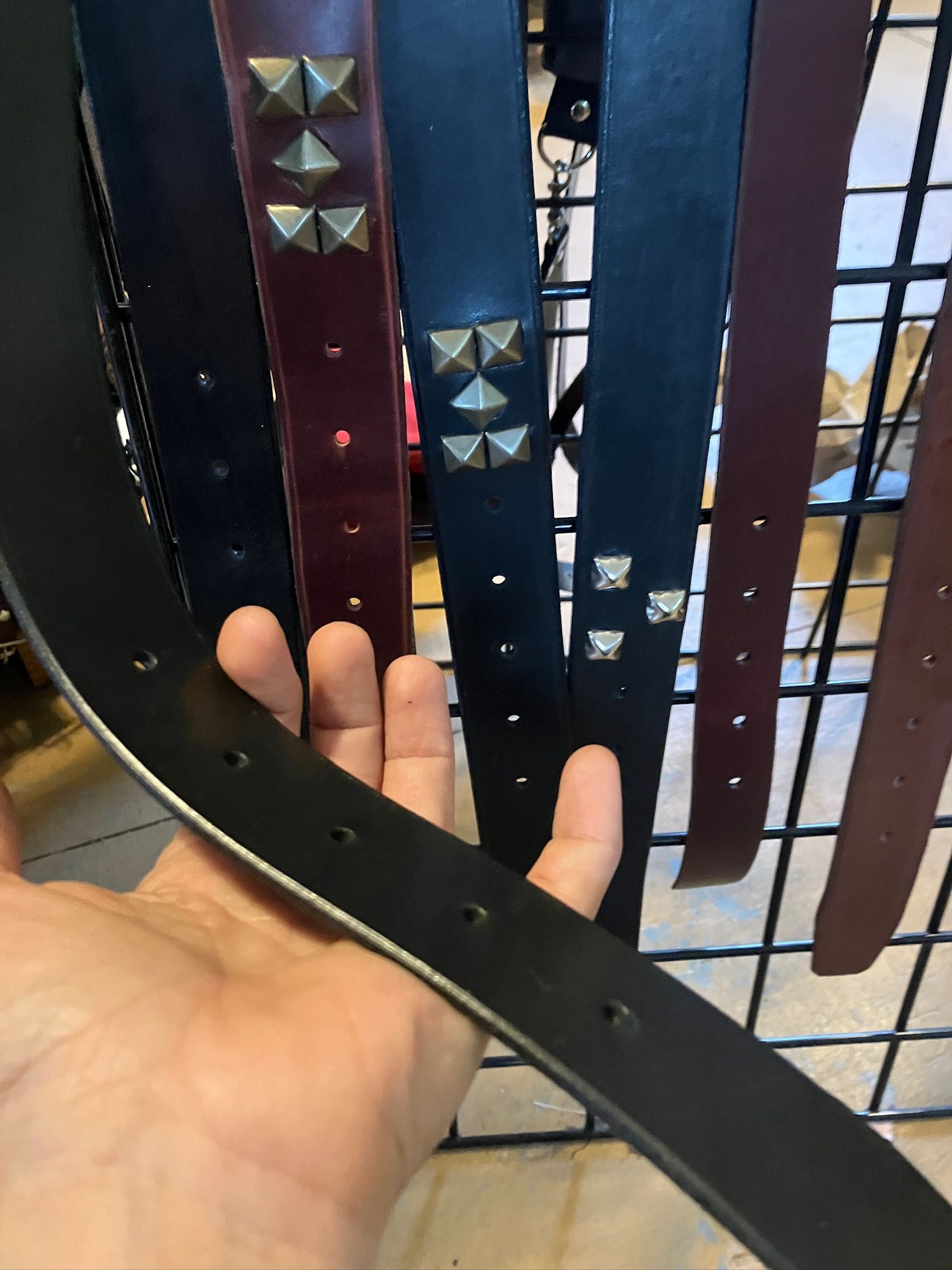 Hand holding black leather belt, several studded belts on display rack.
