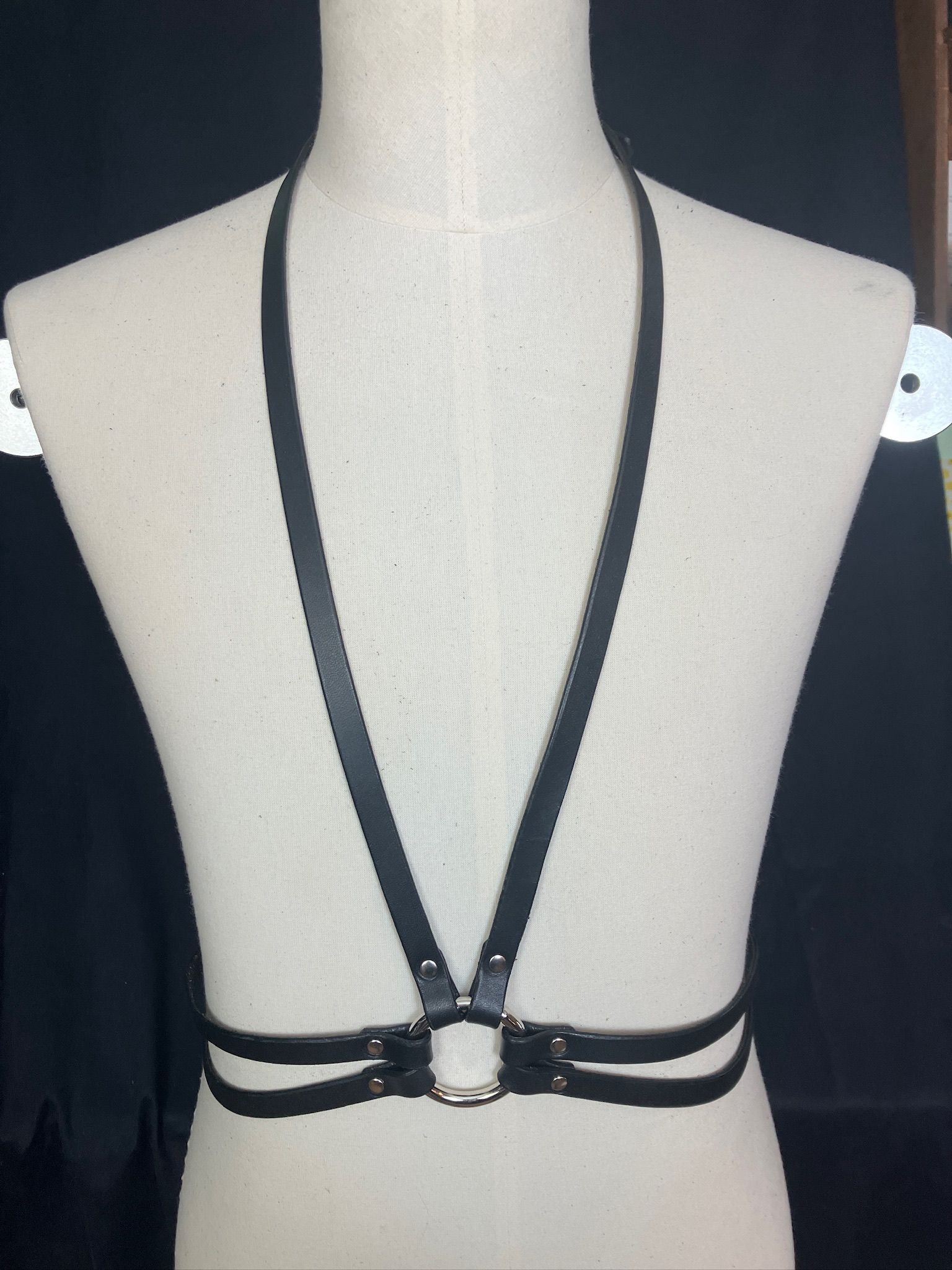Black leather body harness on a white mannequin, with vertical straps over shoulders and horizontal around the torso.
