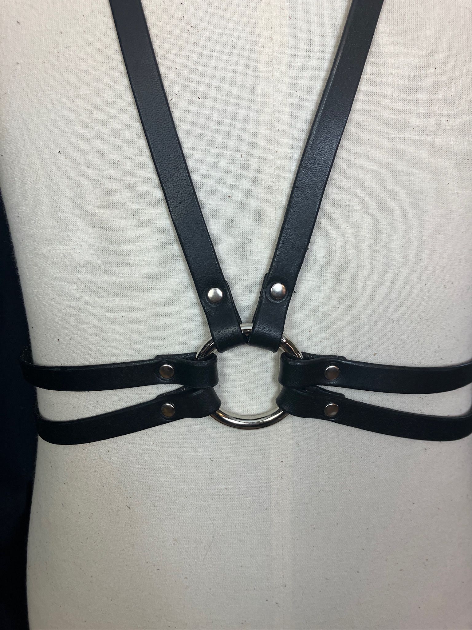 Black leather body harness on a white mannequin, centered on the chest and torso, with silver rings and rivets.