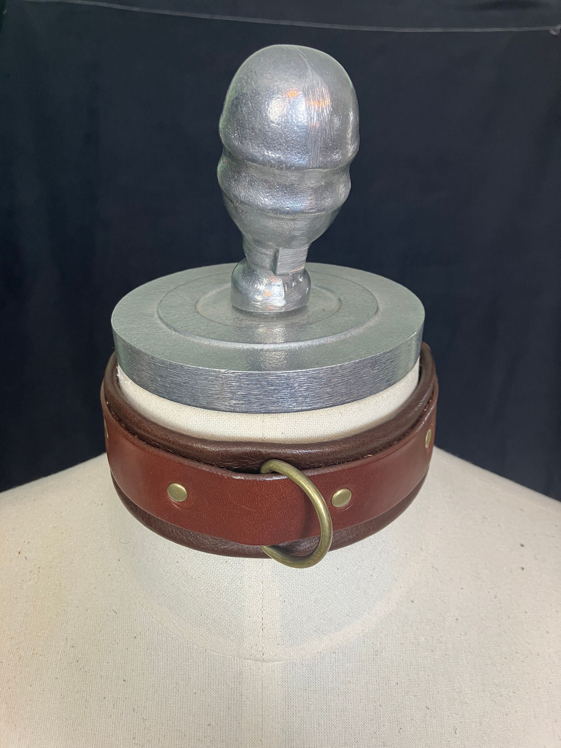 Brown leather collar with brass ring and rivets around a mannequin neck, below a silver head-shaped display.