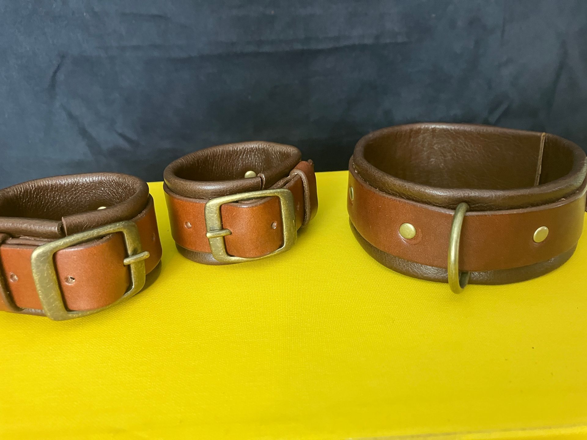 Three brown leather wrist cuffs with gold buckles, sitting on a yellow surface.