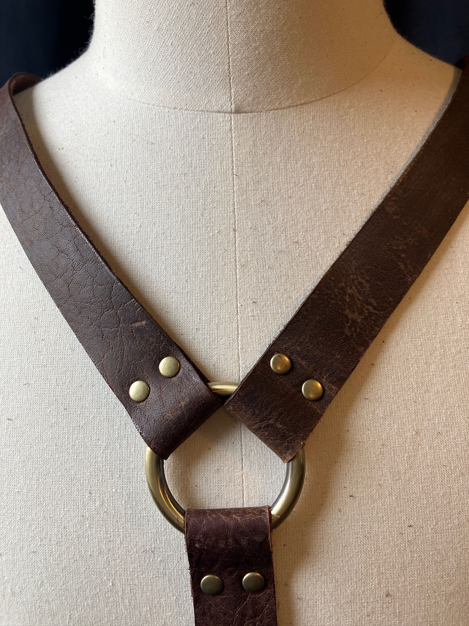 Brown leather harness with metal ring and rivets, on a light-colored torso form.