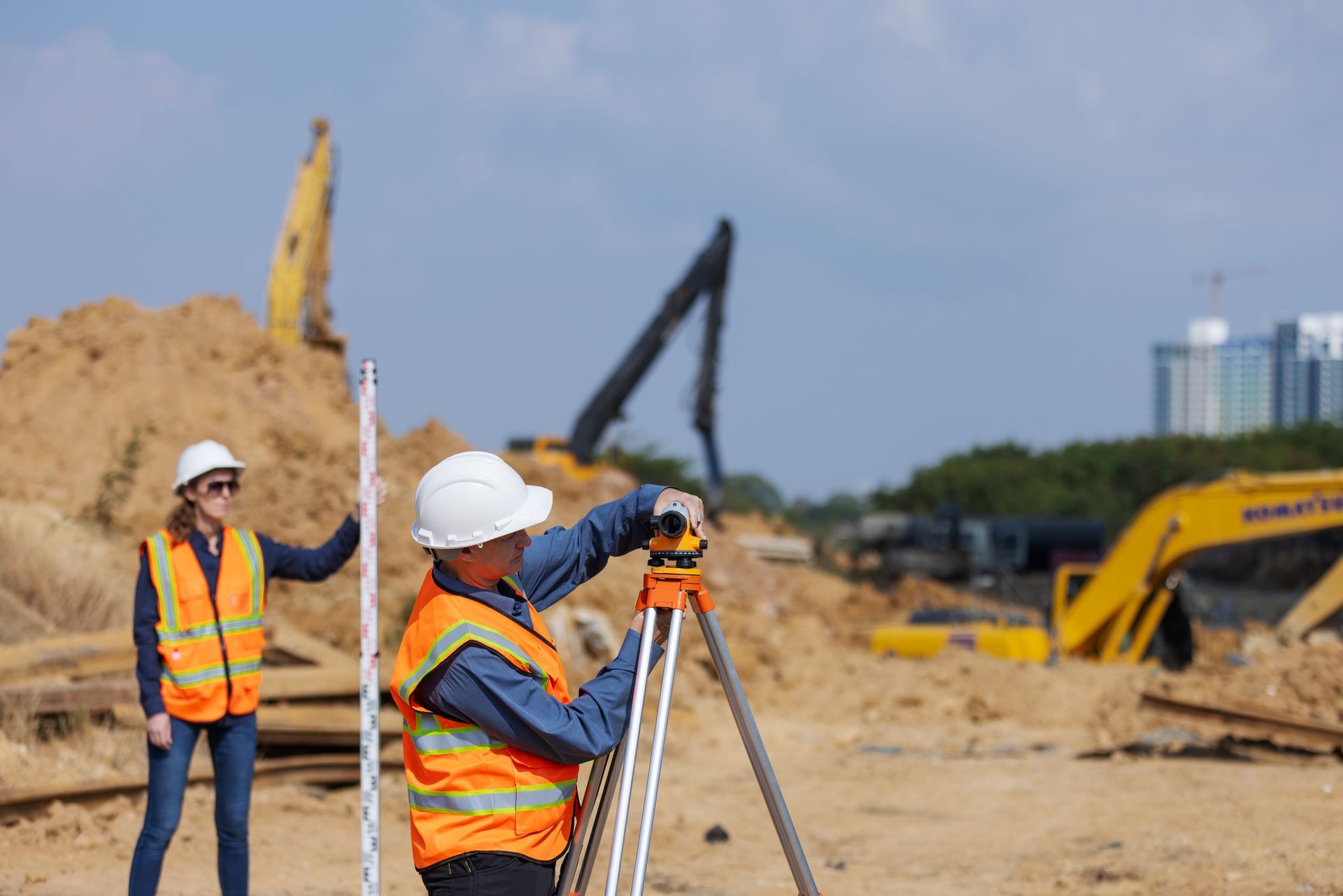 Construction surveyors using a tripod on an active job site with excavators and earthworks underway.