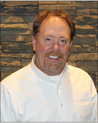A man in a white shirt is smiling in front of a stone wall.