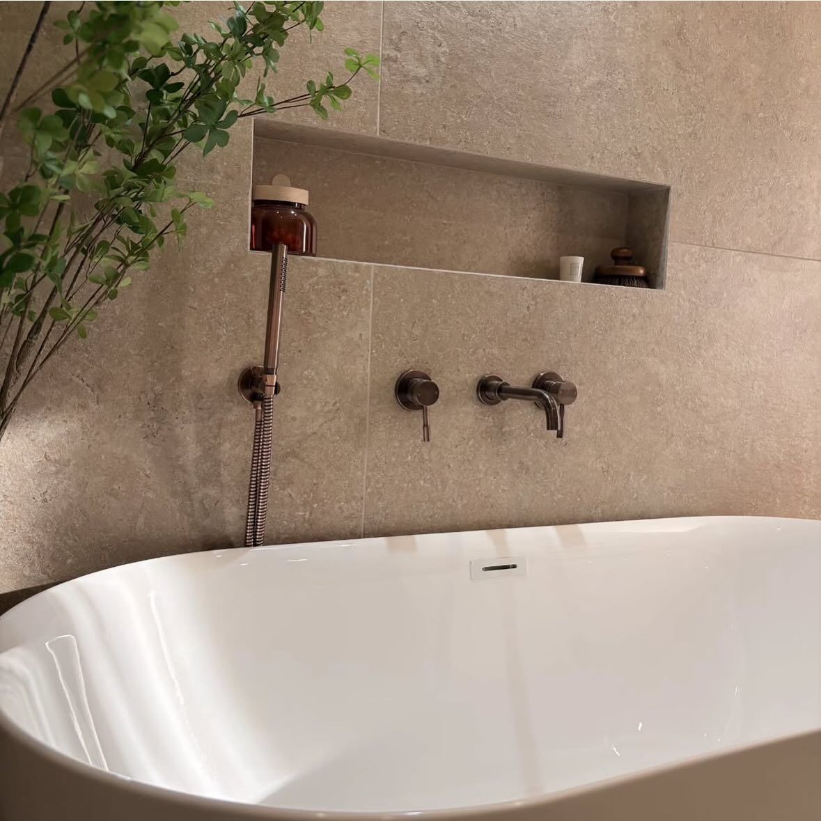 Una vasca da bagno freestanding bianca, posizionata contro una parete rivestita di piastrelle beige con texture, con mensola incassata integrata e rubinetteria in bronzo.