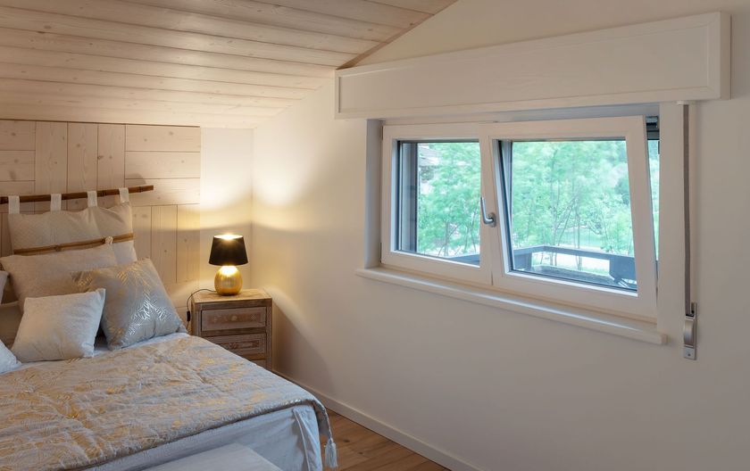 Una camera da letto accogliente con soffitto in legno inclinato, un letto con lenzuola dalla texture particolare, una lampada da comodino e una finestra aperta con vista.