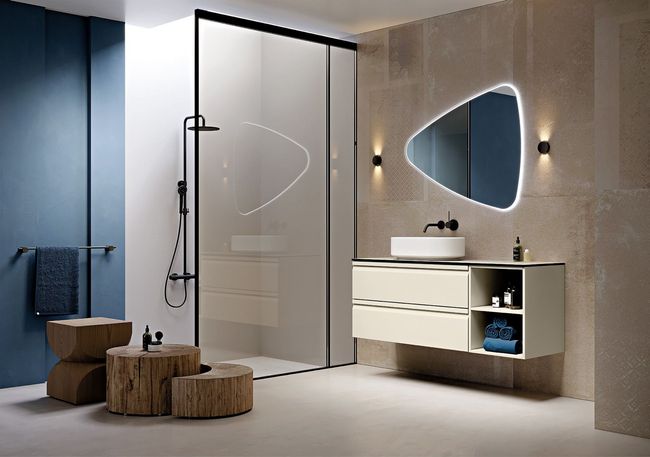 Bagno moderno con parete effetto pietra, mobile lavabo sospeso, specchio triangolare a LED e sgabelli in legno accanto a una cabina doccia in vetro.