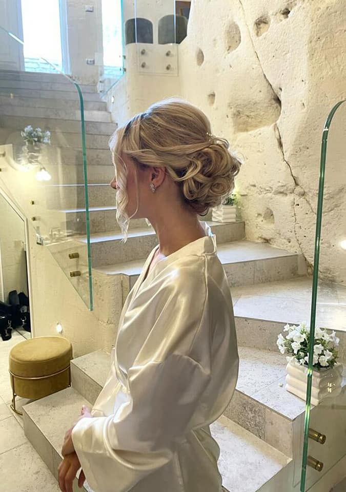 Donna con acconciatura bionda e vestaglia bianca, seduta sulle scale in una stanza luminosa con pareti in pietra.