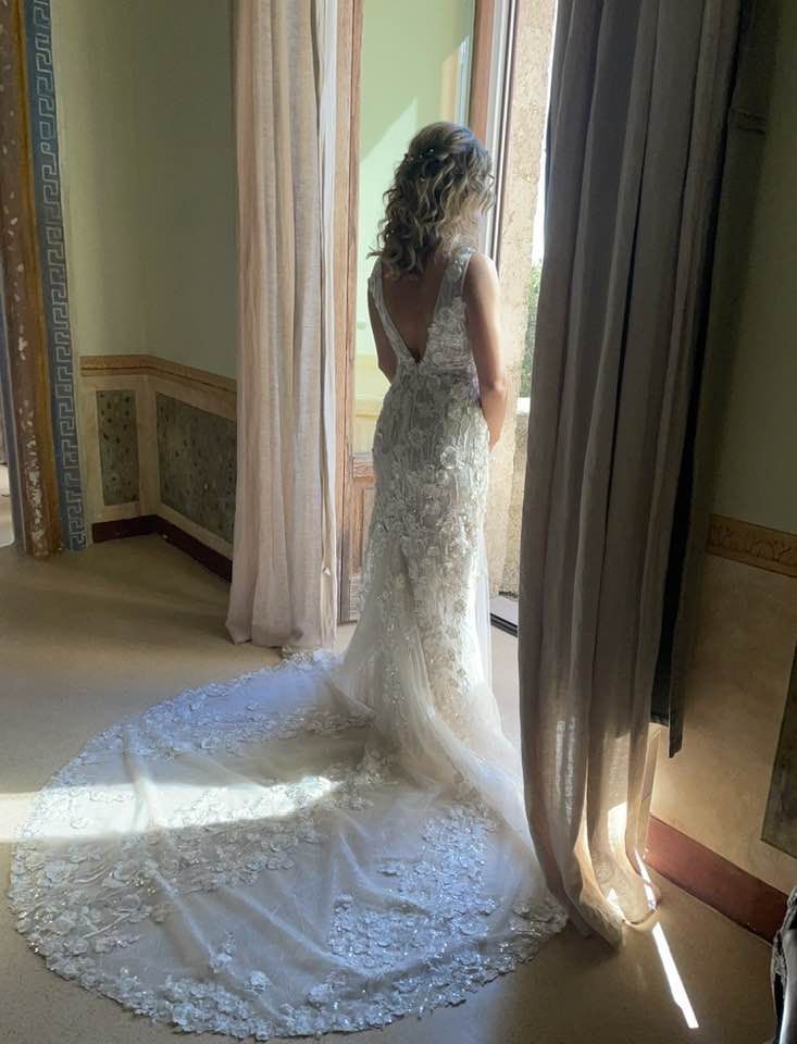 Donna in abito da sposa bianco con lungo strascico, in piedi vicino a una finestra con vista.