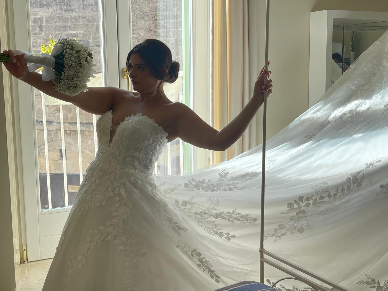 Sposa in abito bianco con in mano un bouquet, in posa vicino a una finestra e a un letto.