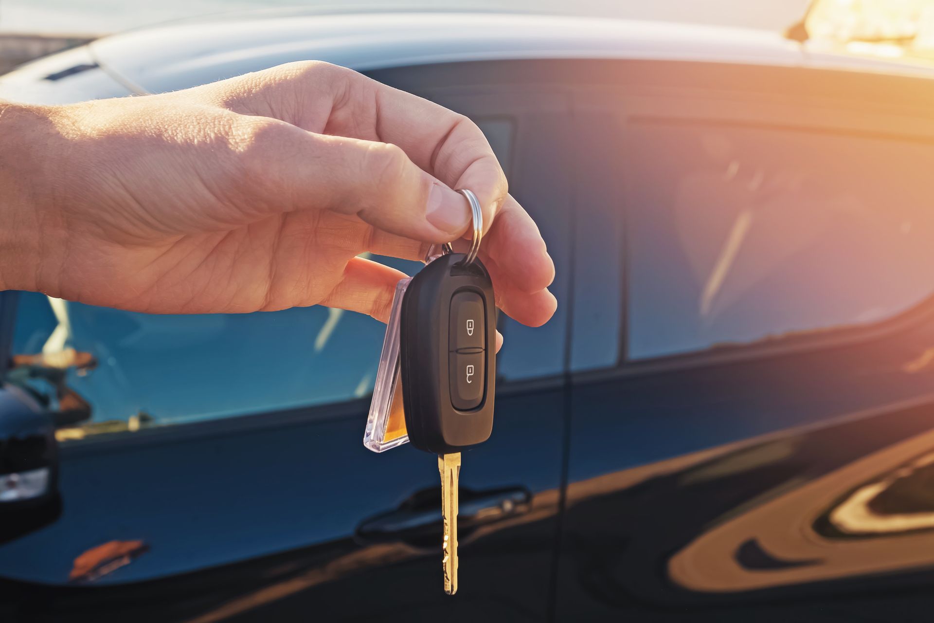 Hand holding car keys in front of a black car, sunlight in the background.
