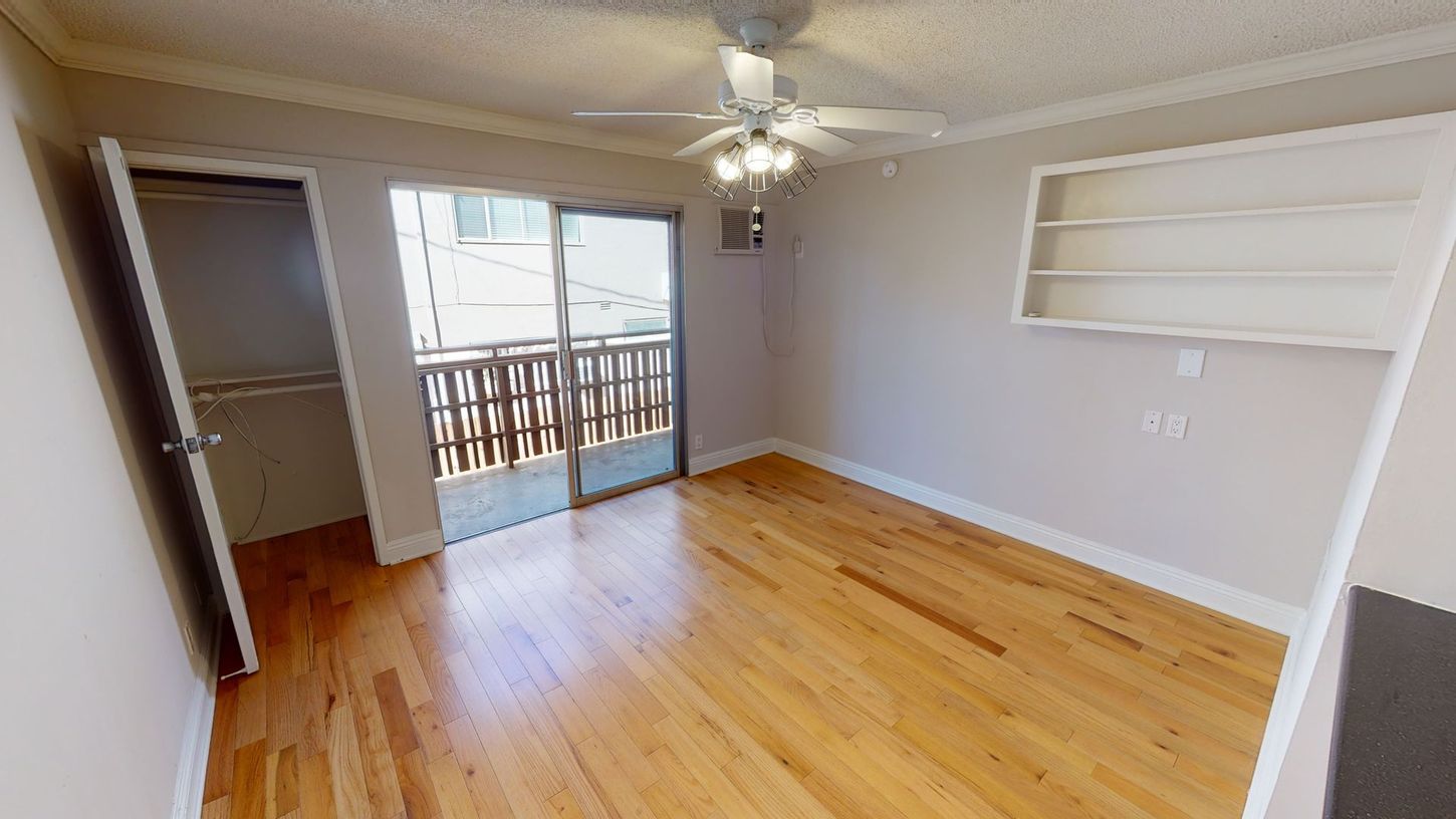 Empty room with hardwood floors, sliding glass door to balcony, built-in shelving, and a ceiling fan.