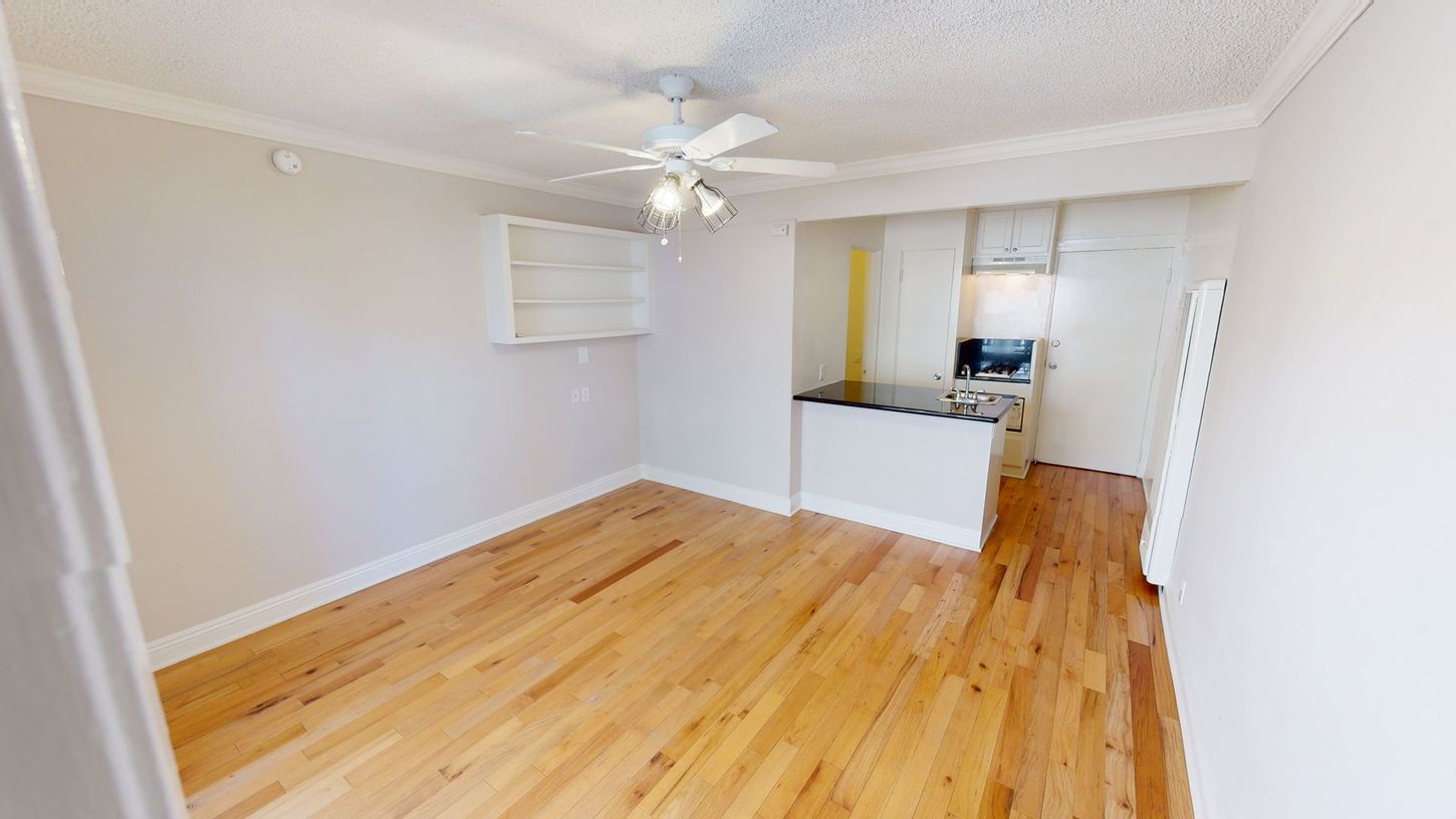 Empty apartment with hardwood floors, a small kitchen, and a ceiling fan.