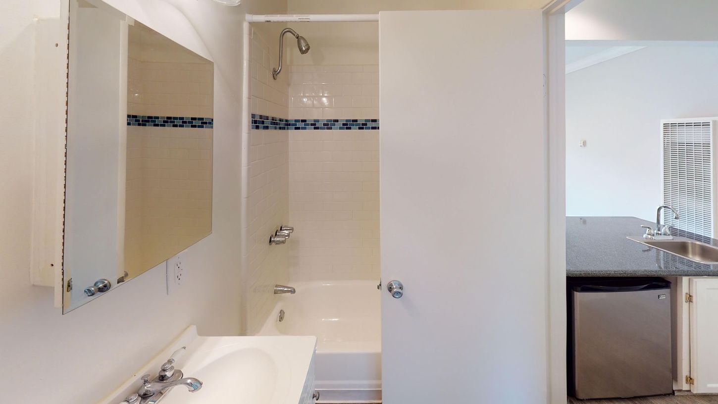 Bathroom with a white door open to reveal a shower, sink, and medicine cabinet; a kitchen is visible in the background.