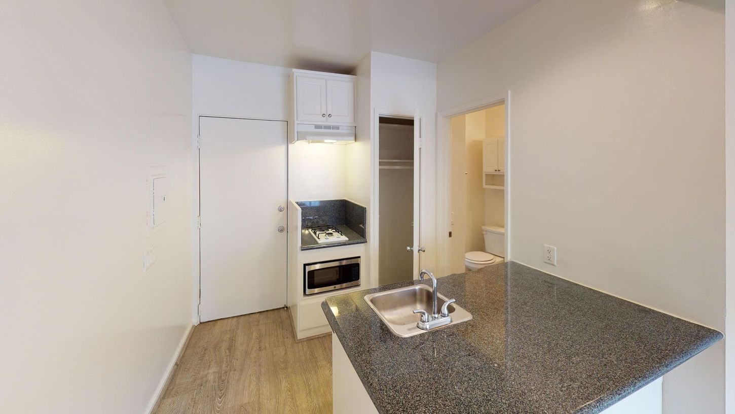 Small apartment kitchen with a countertop, sink, and appliances. A bathroom is in the background.
