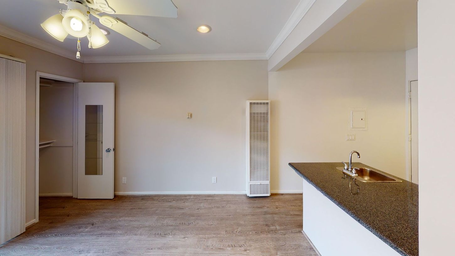 Empty living space with a kitchen counter, closet, heater, and ceiling fan. Light-colored walls and flooring.
