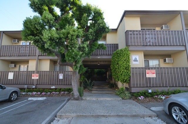 Apartment building with two floors, brown railings, and a large tree in front of the entrance. Cars parked nearby.