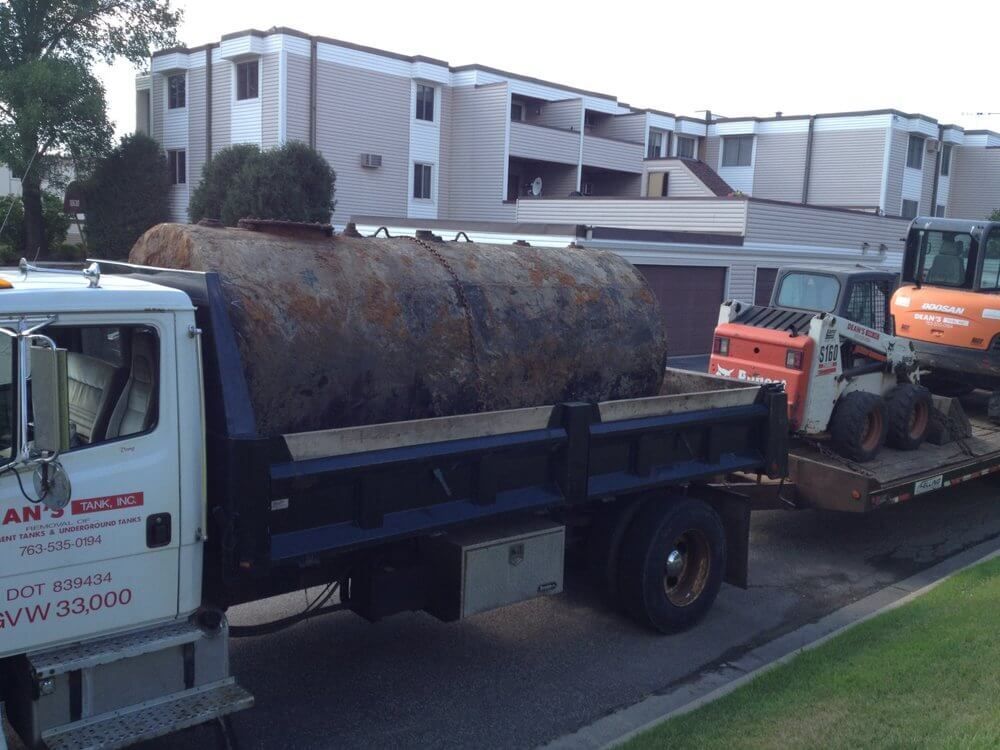 A rusty tank loaded on a truck bed with construction equipment in front of an apartment building.